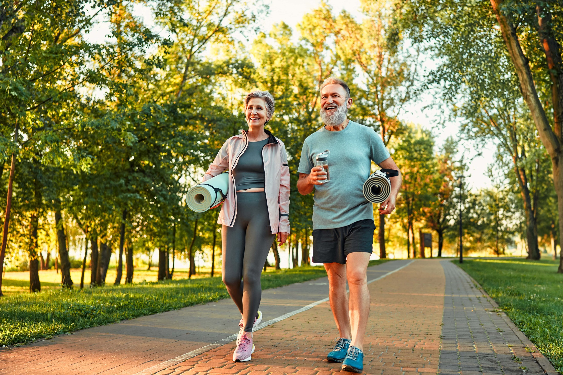 Photo of an older couple walking in a wooded area holding yoga mats